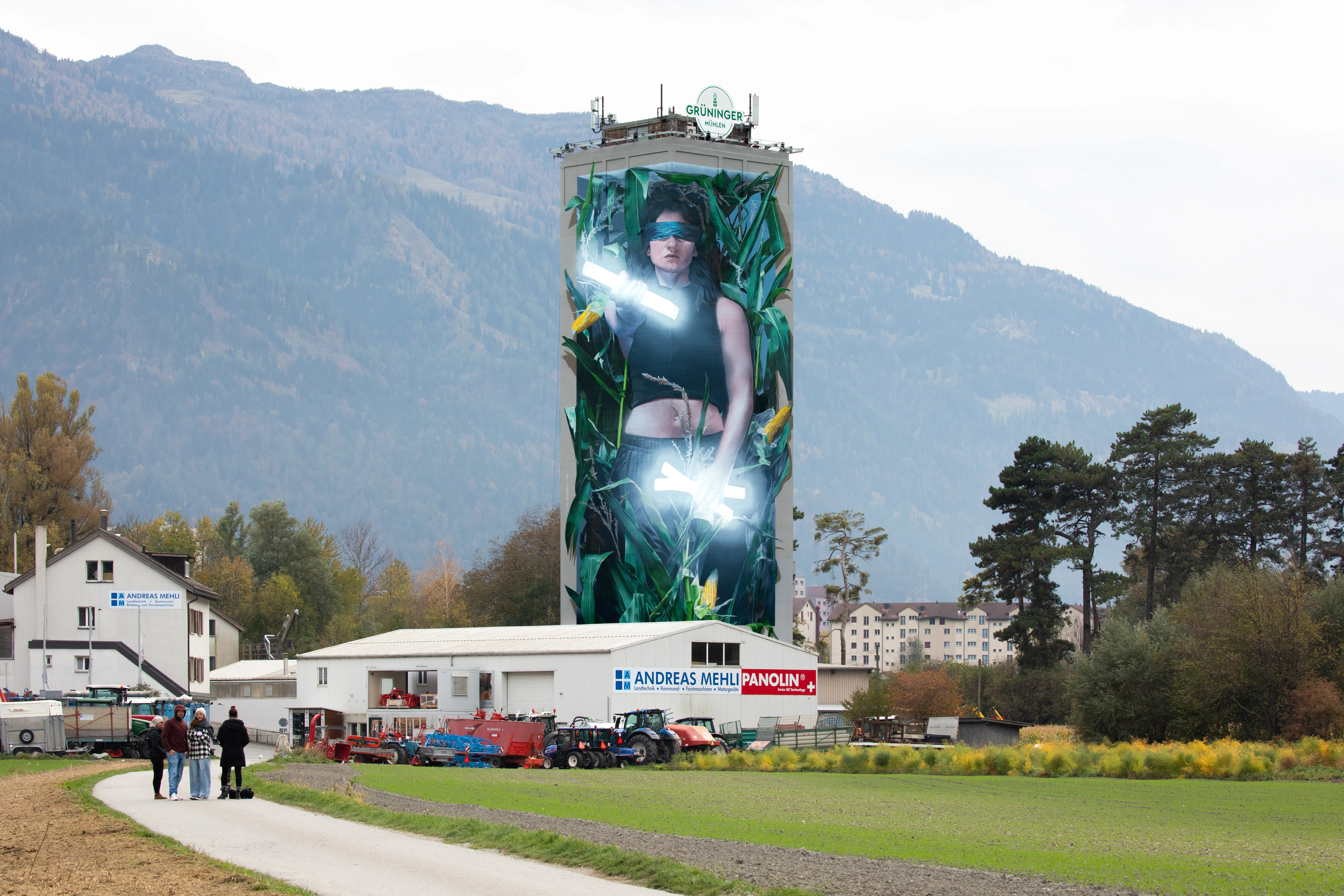 Mühleturm in Chur mit grossem Mural einer Frau zwischen Pflanzen, umgeben von Berglandschaft und ländlicher Umgebung.